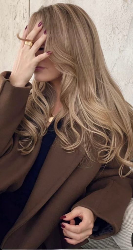 Blondes Haar: Eine Frau mit langen, welligen blonden Haaren trägt einen braunen Mantel.